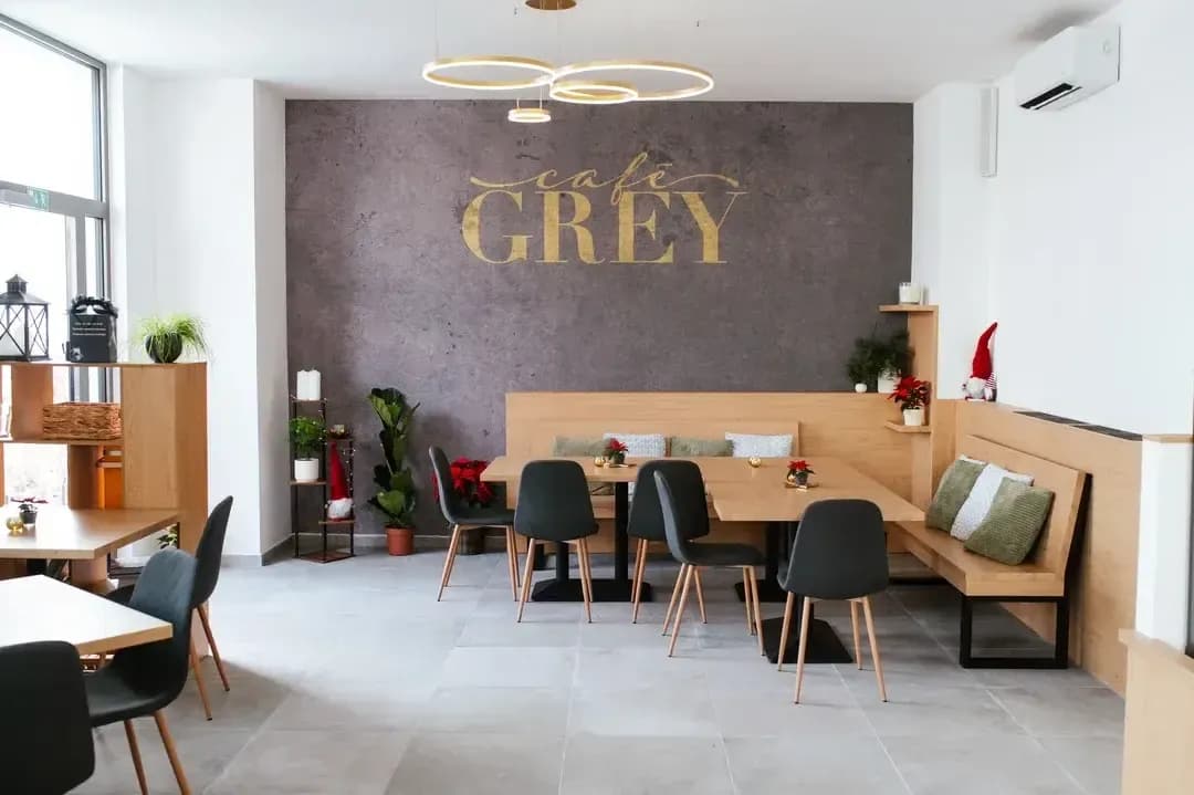 Prostorný interiér Café Grey s dřevěnými lavicemi a stoly, tmavými židlemi a velkým zlatým logem na šedé stěně.