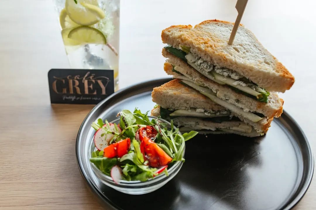 Detail čerstvého sendviče na černém talíři s malým salátem, doplněný o domácí limonádu s limetkou a vizitku kavárny.