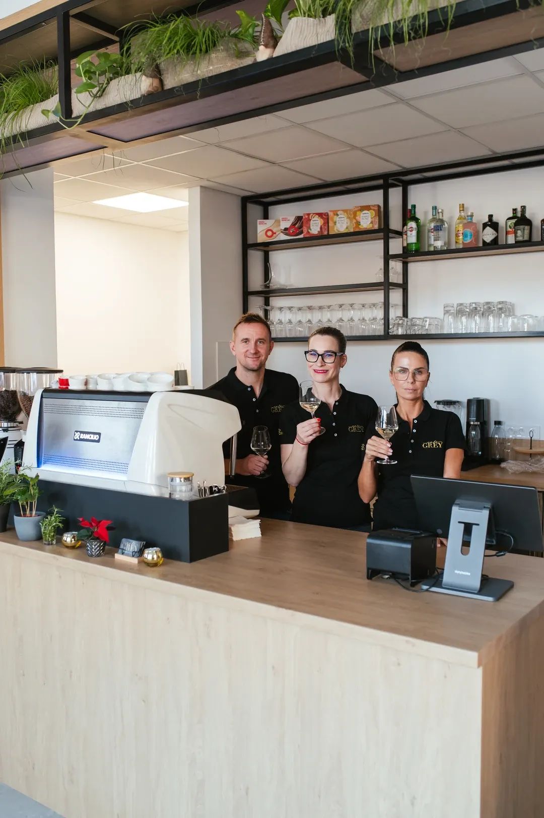 Usměvavý personál kavárny v pracovním oblečení pózující se skleničkami vína za barem s profesionálním kávovarem.