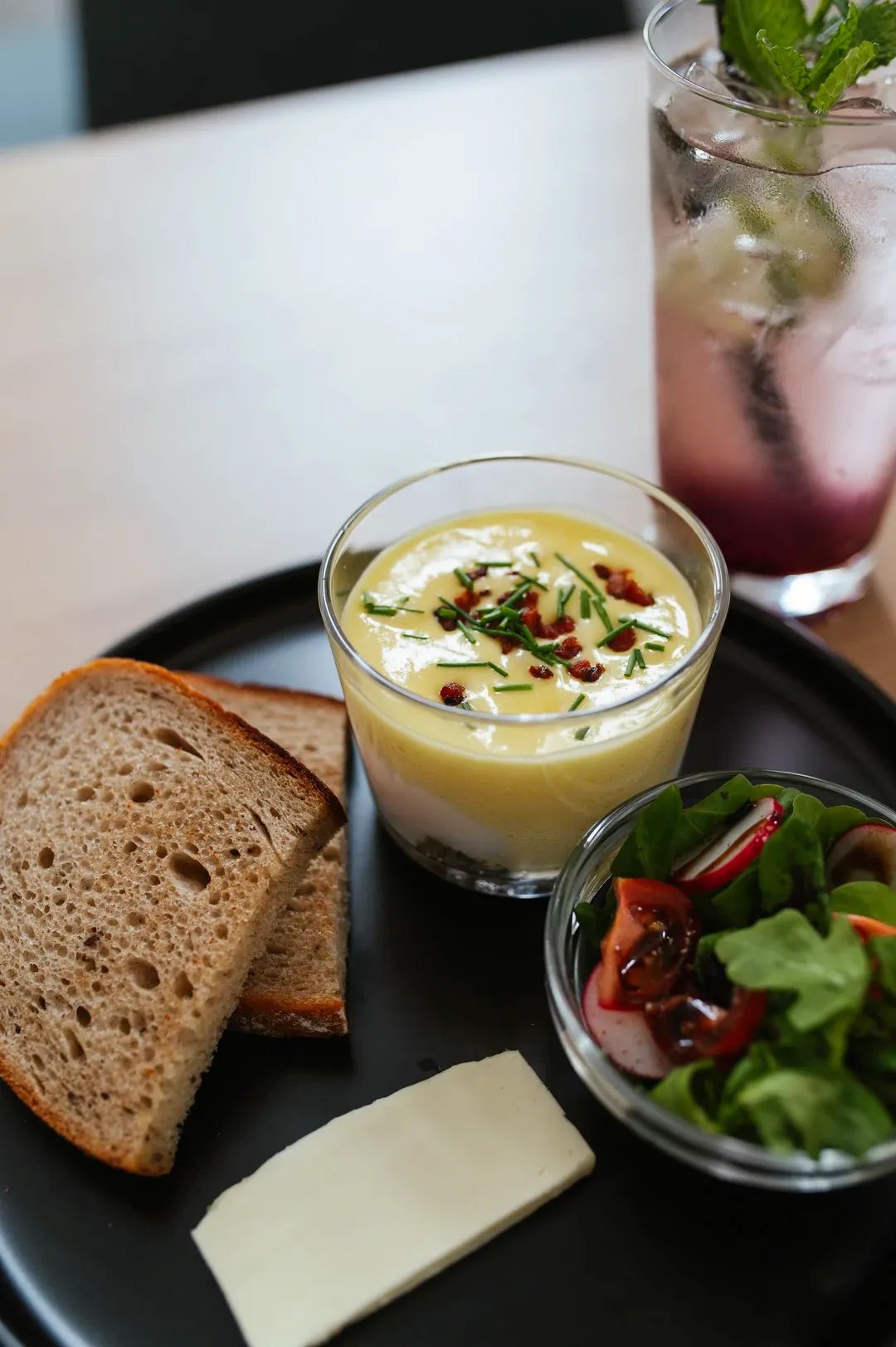 Detail snídaně servírované na černém talíři, zahrnující vajíčkovou pěnu ve skle s pažitkou, chléb, malý salát a růžovou limonádu v pozadí.