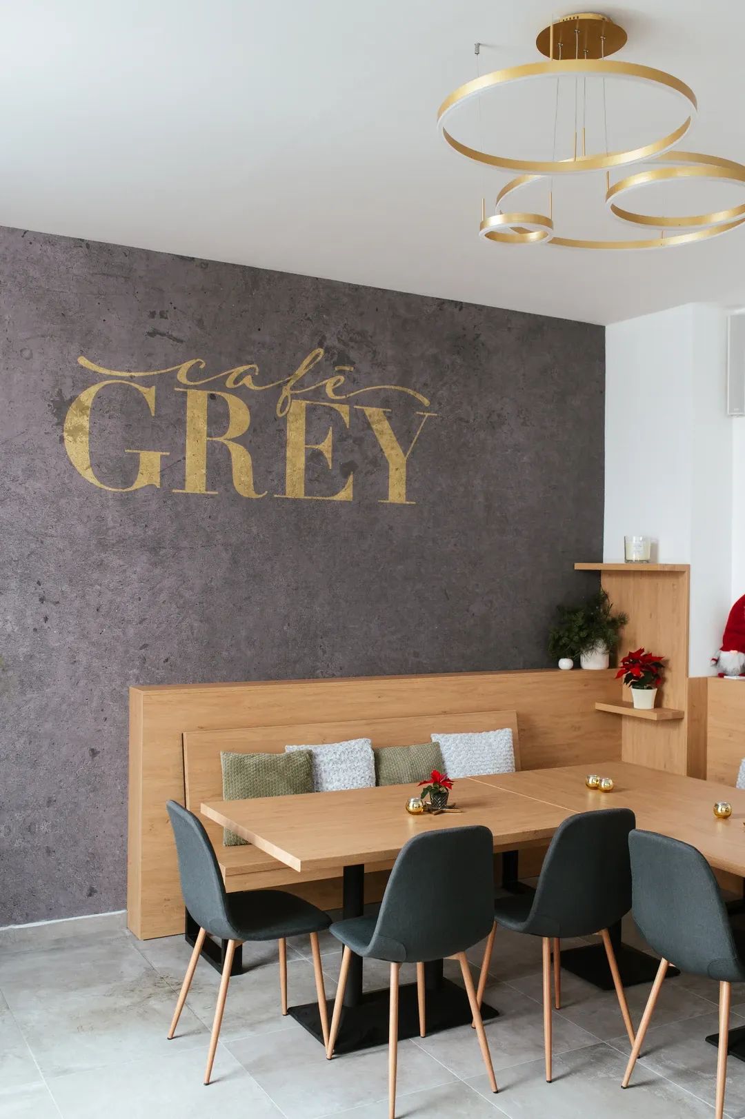 Interiér kavárny s velkým zlatým nápisem Café Grey na tmavě šedé texturované stěně, dřevěným nábytkem a moderním kruhovým lustrem.