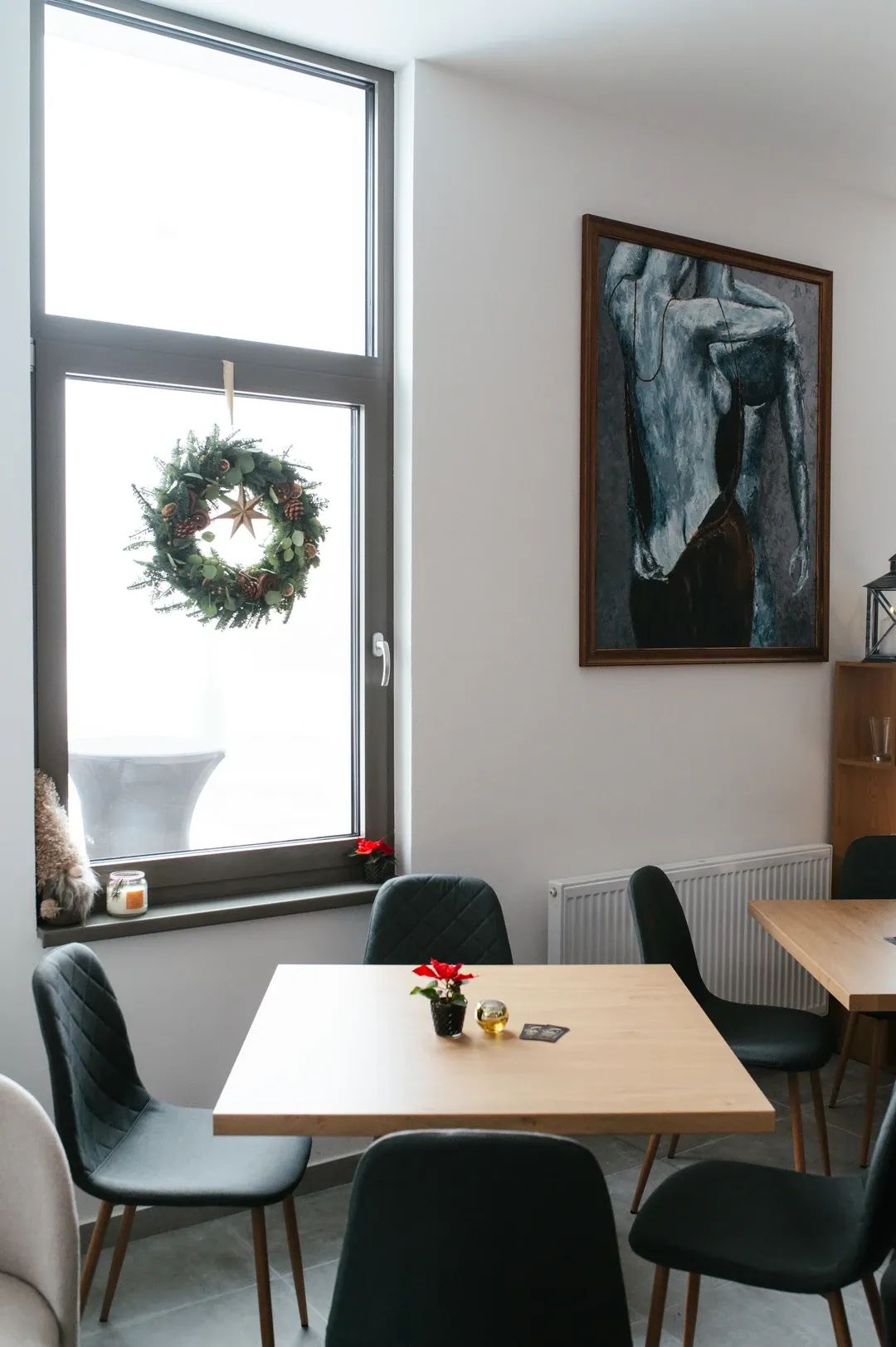 Zákoutí kavárny s dřevěným stolem a černými židlemi u okna s vánočním věncem, na stěně visí obraz postavy v modrých tónech.