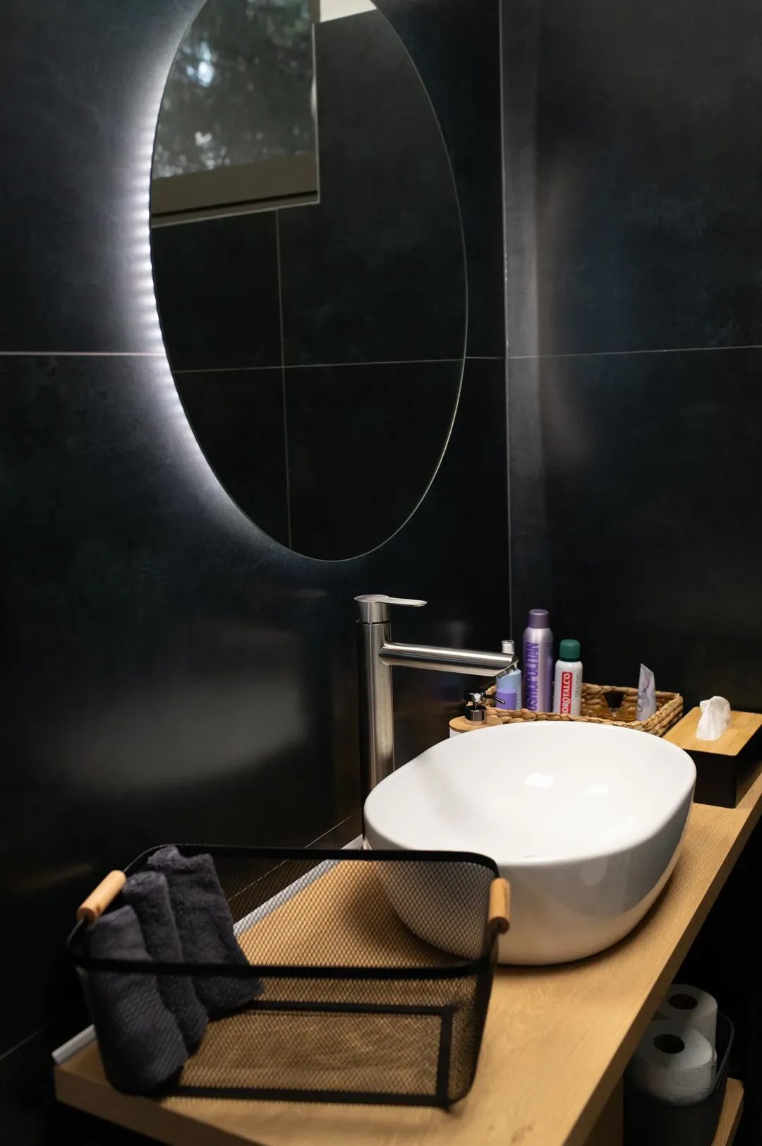 Moderní koupelna s bílým umyvadlem na dřevěné desce, podsvíceným kulatým zrcadlem a tmavým obkladem stěn.