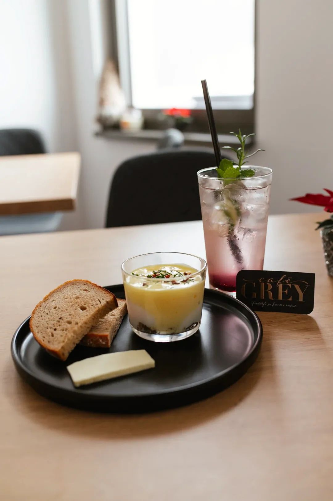 Snídaňový set na stole s vizitkou Café Grey, pokrmem ve skleničce, pečivem a osvěžujícím nápojem s ledem a bylinkami.