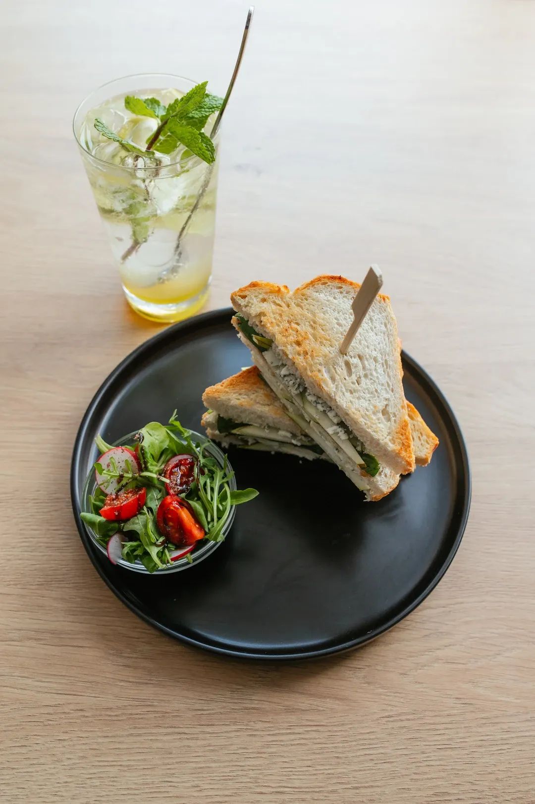 Opečený sendvič z toustového chleba rozkrojený na poloviny, podávaný s čerstvým salátem a domácí limonádou s mátou na dřevěném stole.