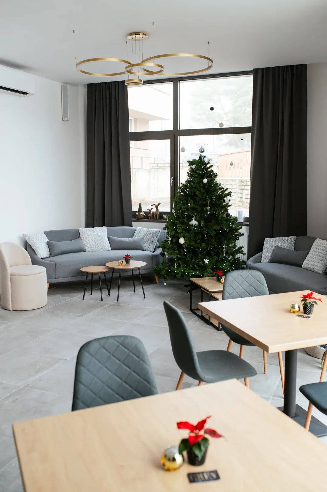 Odpočinková část kavárny s šedými pohovkami, kulatými stolky a ozdobeným vánočním stromečkem u velkého okna.