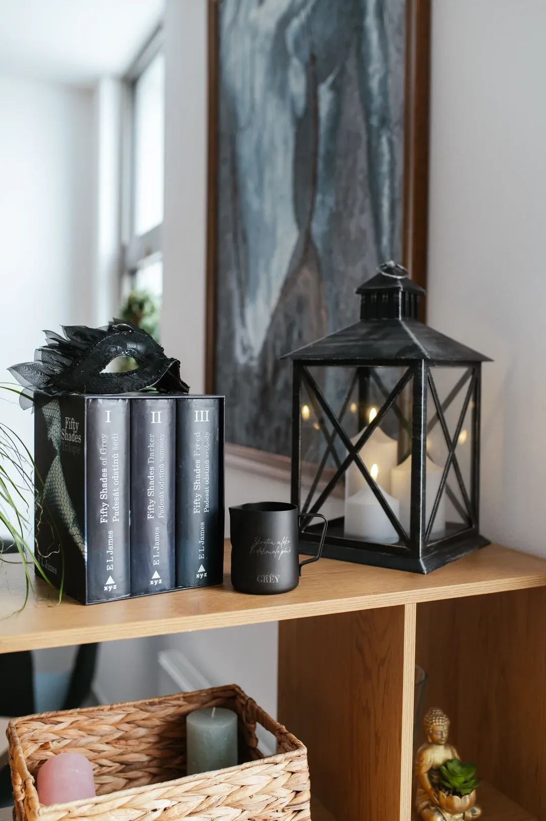 Tematická dekorace na polici s trilogii knih Padesát odstínů šedi, černou škraboškou, hrnkem a velkou lucernou se svíčkami.