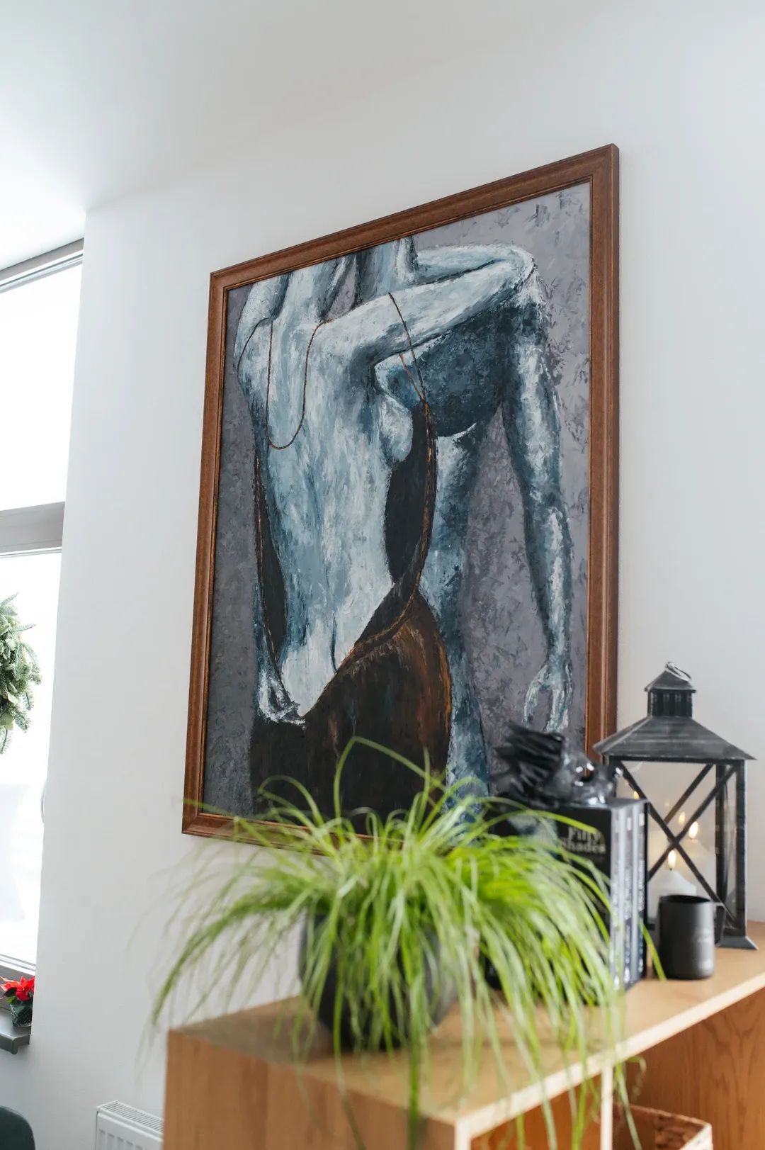 Detail uměleckého obrazu na zdi v modro-šedých barvách, v popředí zelená pokojová rostlina a dekorativní lucerna.