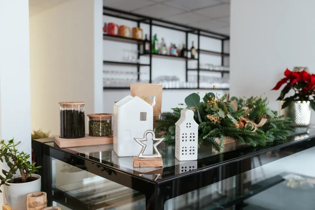 Pult v kavárně s vánoční výzdobou, bílými keramickými domečky, jehličím a sklenicemi se sypaným čajem.