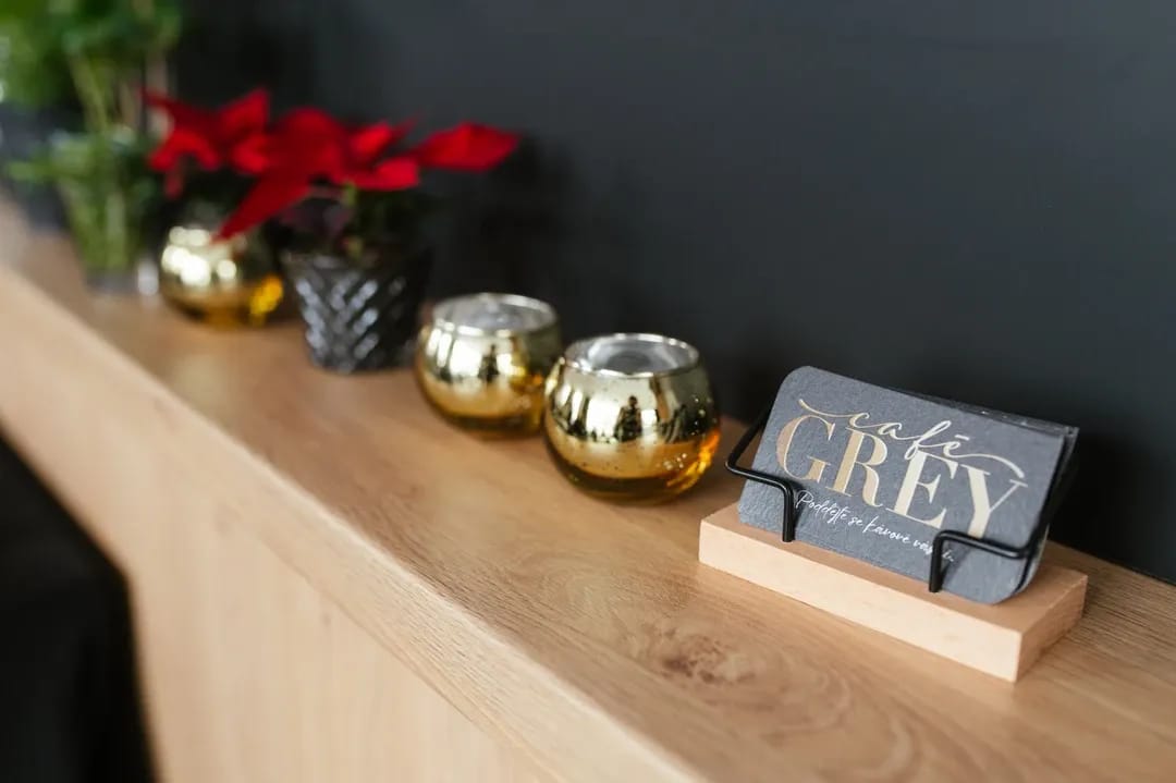 Detail černých vizitek se zlatým logem Café Grey umístěných ve stojánku na dřevěné polici vedle zlatých svícnů.